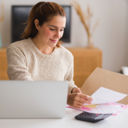 Smiling woman sitting at desk in front of laptop handling paperwork inside of a folder with calculator in front